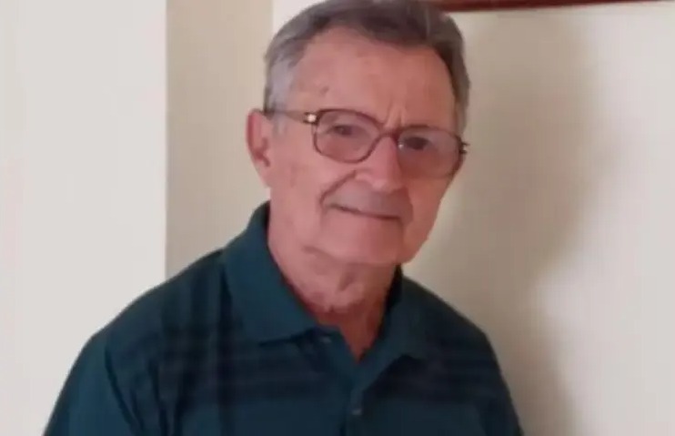 Homem idoso de óculos, vestindo camisa polo escura, em ambiente interno, com expressão neutra.