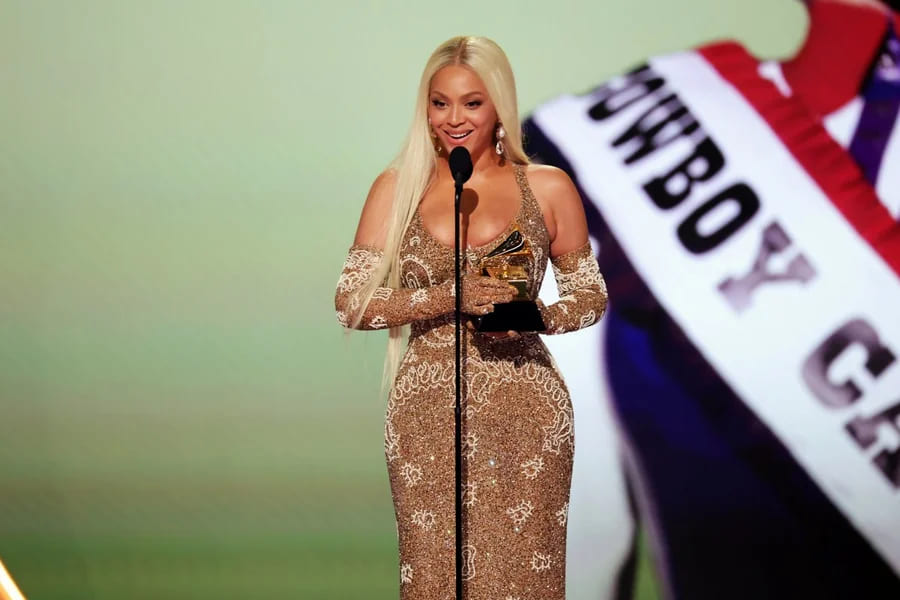 Beyonce com vestido dourado brilhante e luvas longas, segurando um prêmio e discursando em um microfone no palco, com fundo desfocado e faixa ao fundo.